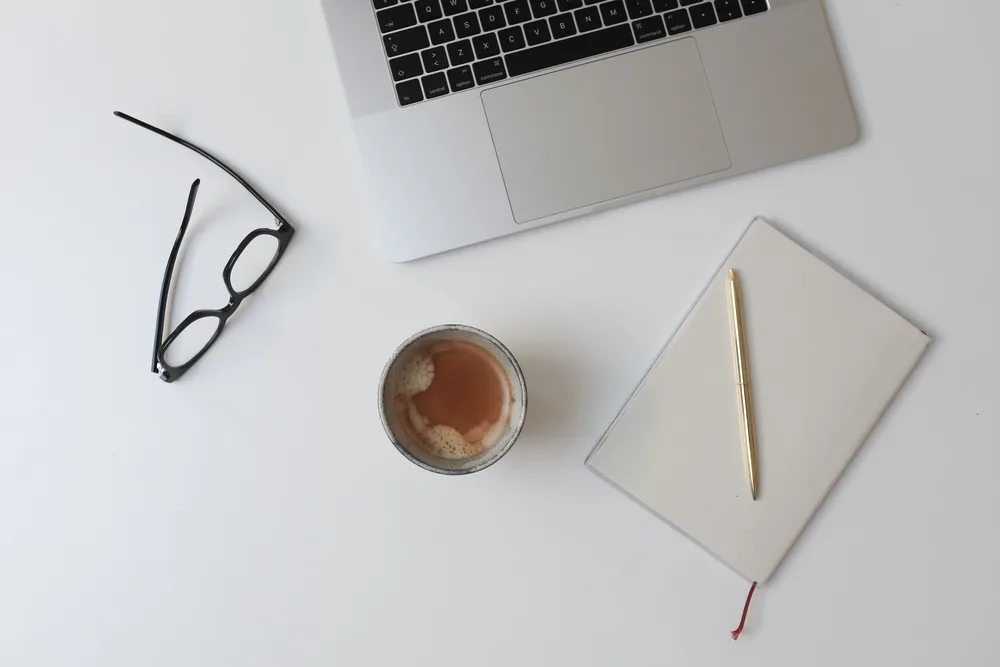 Laptop, Notizbuch und Brille auf einem hellen Schreibtisch